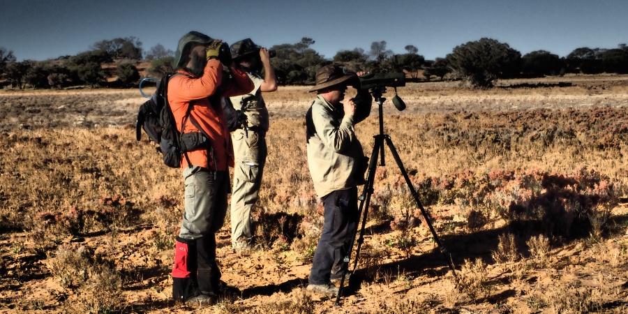 Three researchers looking through binoculars and telescopes standing in a bare landscape